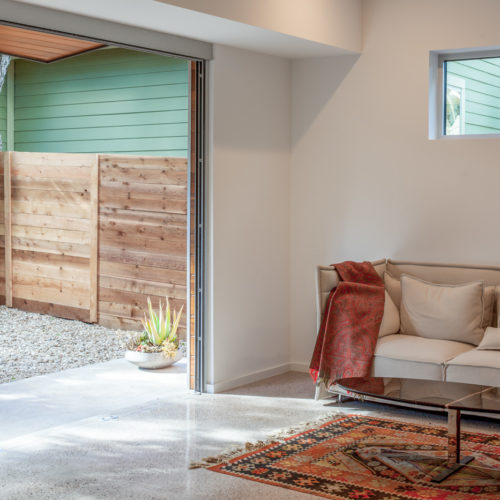 A couch and coffee table inside connected directly to a backyard through a large garage door sized opening in a wall.