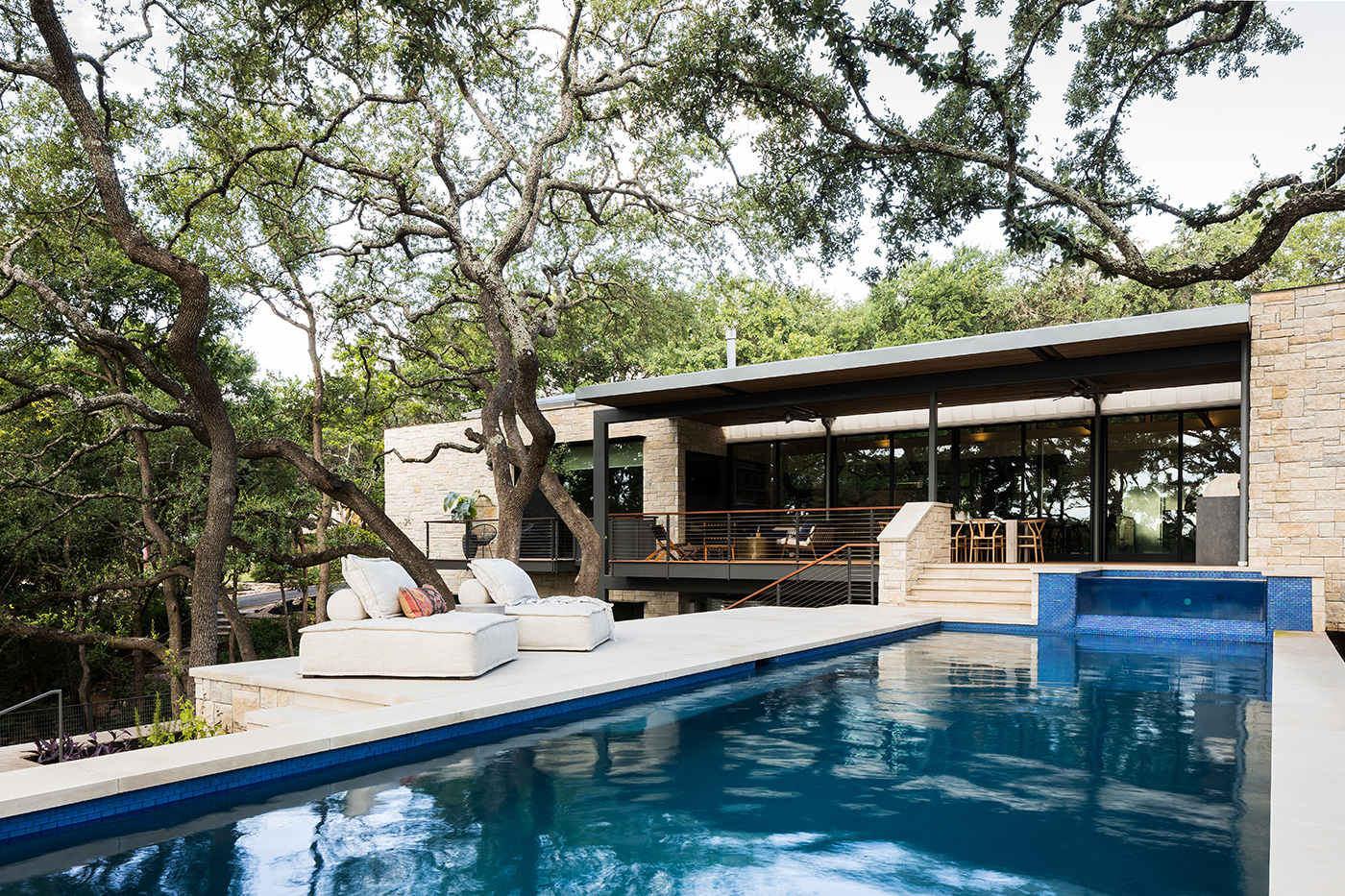 The tree covered pool and back patio of a house.