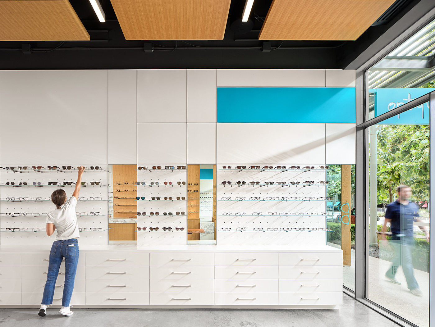 A girl reaching up to grab glasses from the top shelf of a glasses store.