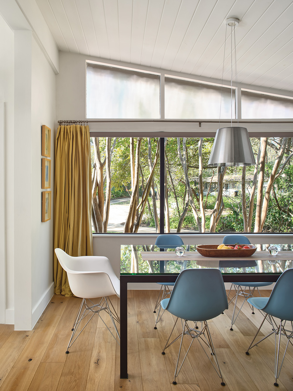 Sleek dining room with glass table and large window that show front yard landscaping.