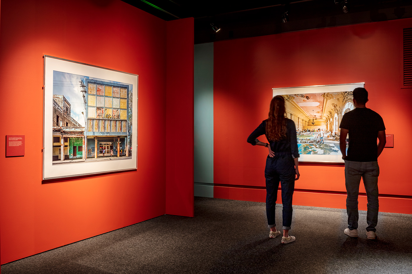 Two people looking at painting on a red exhibit wall.