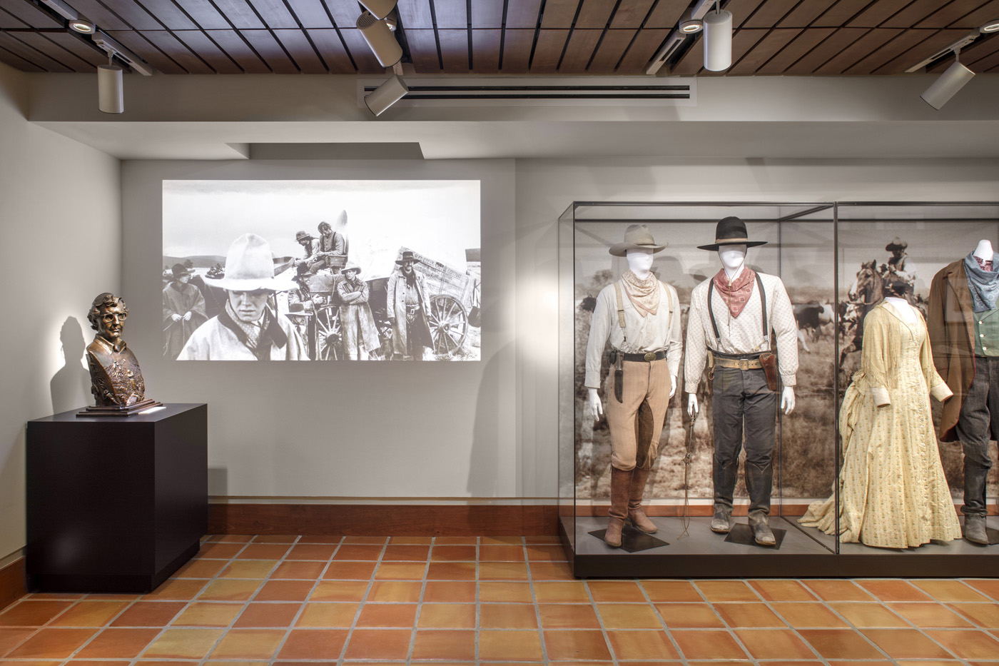 Maniquis in western attire behind glass display cases with a black and white film being displayed on wall.