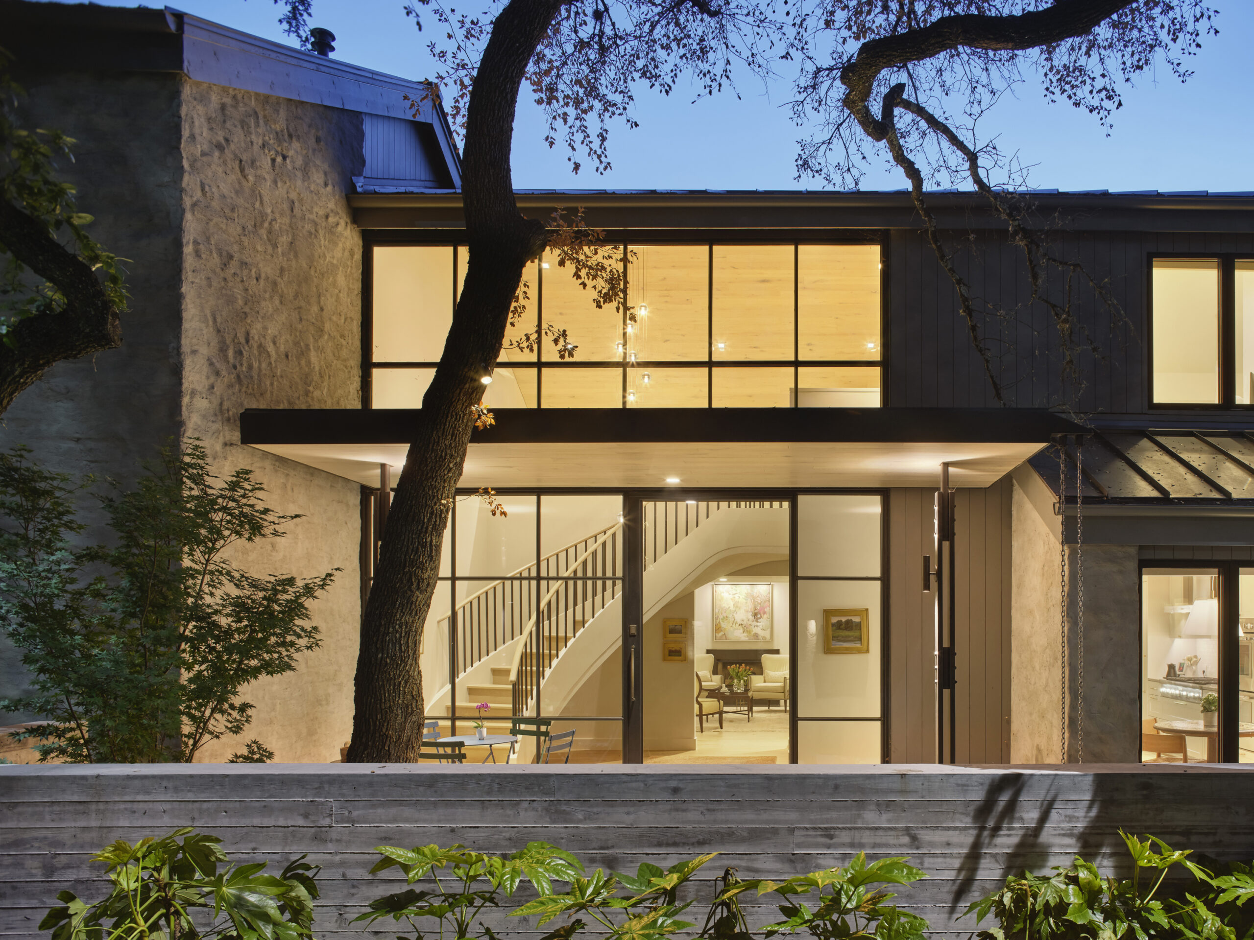 residence exterior at dusk with lighted interior looking through glass windows into foyer with winding stair