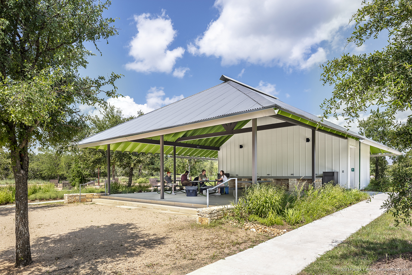 People sitting at picnic tables in a covered outdoor seating area surrounded by trees and landscaping.