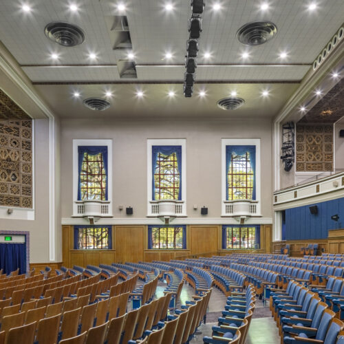 View along curved rows of seats in an auditorium, facing a series of windows.