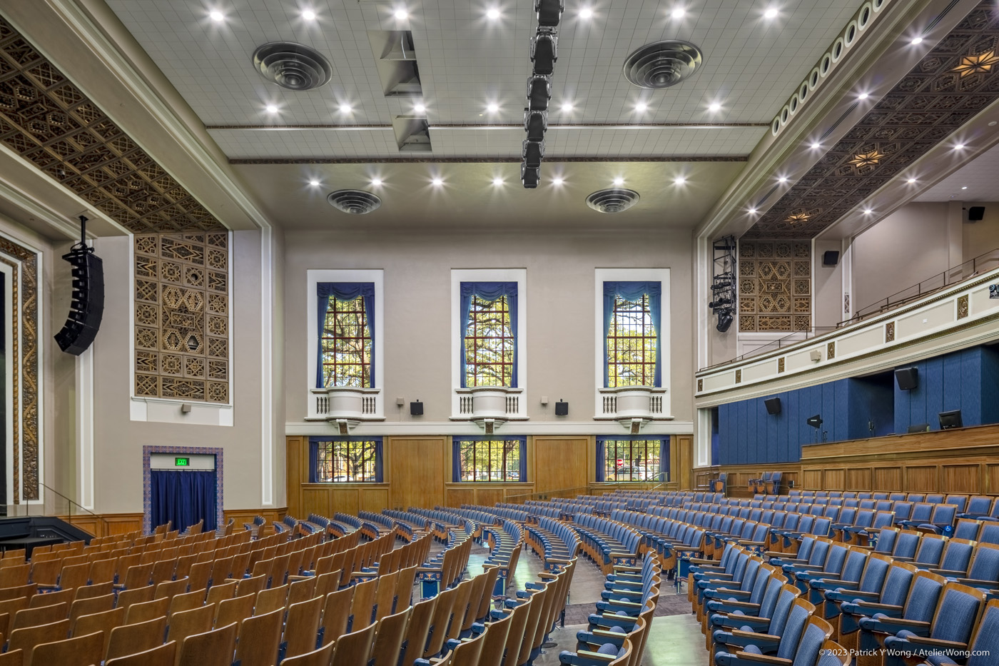 View along curved rows of seats in an auditorium, facing a series of windows.